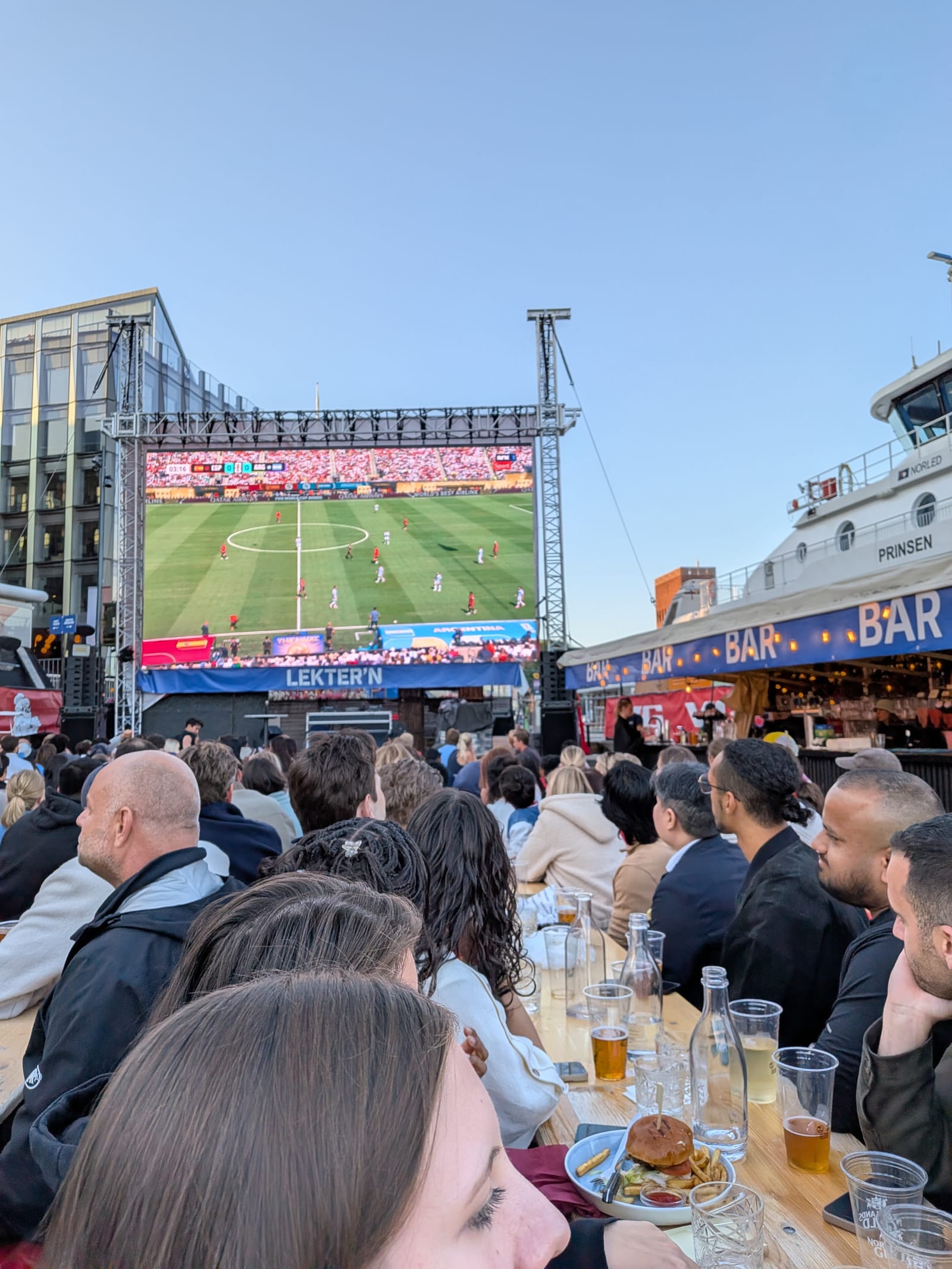 The group watching the match together on the outdoor big screen, tables full, Oslo waterfront in the evening light